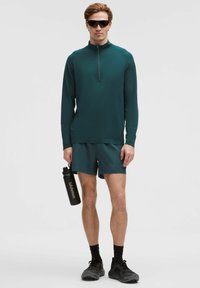 Vyras, dėvintis tamsiai žalią sportinę ilgomis rankovėmis viršutinę dalį ir šortus, laiko juodą Lululemon vandens butelį, su juodomis kojinėmis, sportbačiais ir akinių nuo saulės.