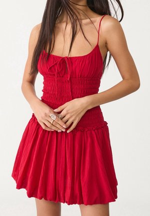 Femme portant une robe rouge sans manches avec de fines bretelles et un corsage froncé, mains jointes devant, portant plusieurs bagues.