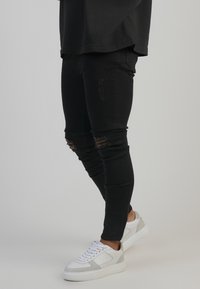 Schwarze, distressed Skinny Jeans mit Rissen an den Knien und einer figurnahen Silhouette. Kombiniert mit weißen Sneakers, die einen strukturierten grauen Akzent zeigen.