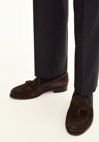 Braune Wildleder-Lederschuhe mit Quastenverzierung und gestickten Akzenten, kombiniert mit dunklen Hosen. Nahaufnahme des Schuhwerks vor einem hellen Hintergrund.
