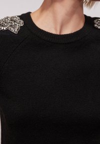 Pull en maille noire avec encolure ronde, tissu texturé et ornements décoratifs en perles sur les deux épaules.