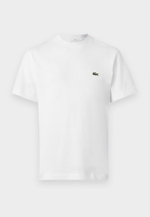 Mann und Frau stehen nebeneinander in weißen Lacoste-T-Shirts und dunkelblauen Lacoste-Sweatpants mit weißen Turnschuhen vor einem schlichten Hintergrund.