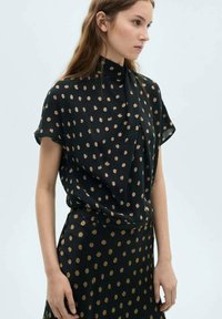 Schwarze Bluse mit kurzen Ärmeln, hohen Kragen und goldenem Punktmuster. Hergestellt aus einem weichen, leichten Stoff.