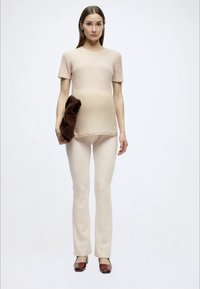 Ανοιχτό μπεζ κοντομάνικη μπλούζα και ταιριαστά στενά παντελόνια από απαλό πλεκτό ύφασμα. Το μοντέλο κρατάει καφέ τσάντα χειρός και φοράει μπορντό παπούτσια.
