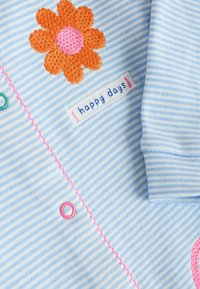 Blauwe gestreepte stof met een oranje geborduurde bloem met een roze midden, en een label met de tekst "gelukkige dagen" in het blauw met roze accenten.