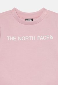 T-shirt di cotone rosa con scollo rotondo, dettagli con bottoni a pressione e testo stampato in bianco "THE NORTH FACE" sulla parte anteriore.