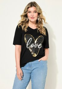 Svart t-shirt med korta ärmar som har en grafik av ett guld- och vitt hjärta med texten "love". Matchad med ljusblå jeans prydda med vita pärlor.
