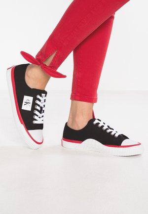 Pieds portant des baskets noires et blanches à lacets avec semelles rouges associés à un pantalon rouge noué en nœud à la cheville sur fond blanc.