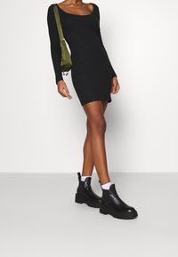 Czarna, prążkowana sukienka z długim rękawem i okrągłym dekoltem, zestawiona z masywnymi czarnymi botkami za kostkę i białymi skarpetkami do kostek. Zielona torebka na ramię jako dodatek.