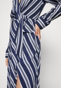 Námořnicky modré a bílé pruhované šaty s vázáním v pase a dlouhými rukávy, s hlubokým výstřihem do V a bočním rozparkem. Hladká látka s jemným leskem.