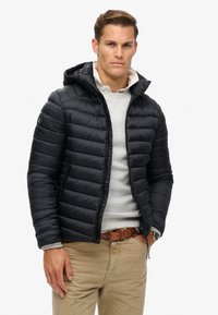 Chaqueta de plumas negra con capucha, que presenta costuras horizontales, cierre de cremallera y bolsillos laterales. Llevada sobre un suéter gris claro.