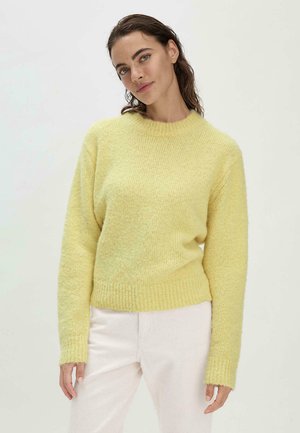 Pull en tricot jaune, moelleux, avec un col rond, un ourlet côtelé et des manches longues, associé à un pantalon de couleur claire. Design minimaliste.