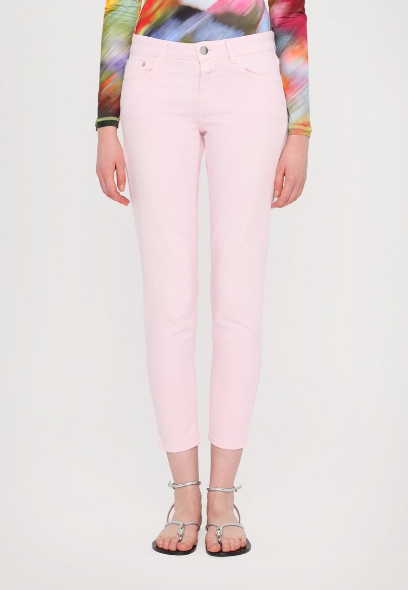 Femme portant un pantalon slim rose clair longueur cheville avec des sandales plates argentées et un haut à manches longues multicolore.