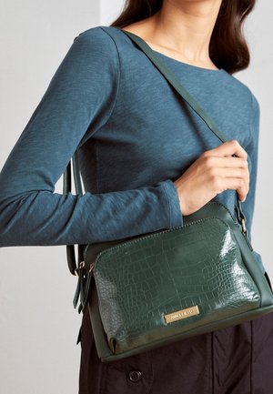 Vrouw draagt een teal top met lange mouwen en houdt een donkere groene, getextureerde schoudertas met gouden rits en een logo op het voorvak vast.