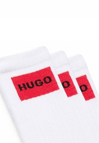 Weiße Socken mit einem roten rechteckigen Band, auf dem die Marke "HUGO" in schwarzer Schrift zu sehen ist. Das Material wirkt weich und dehnbar.