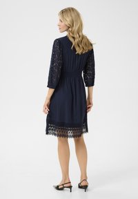 Navy blue dress with three-quarter lace sleeves, a cinched waist, and a lace hem featuring intricate patterns and fringe detailing.