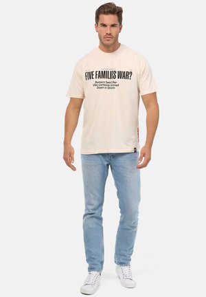 Beige bomulds t-shirt med sort tekst, rund hals og korte ærmer. Sammen med lyseblå jeans og hvide sneakers.