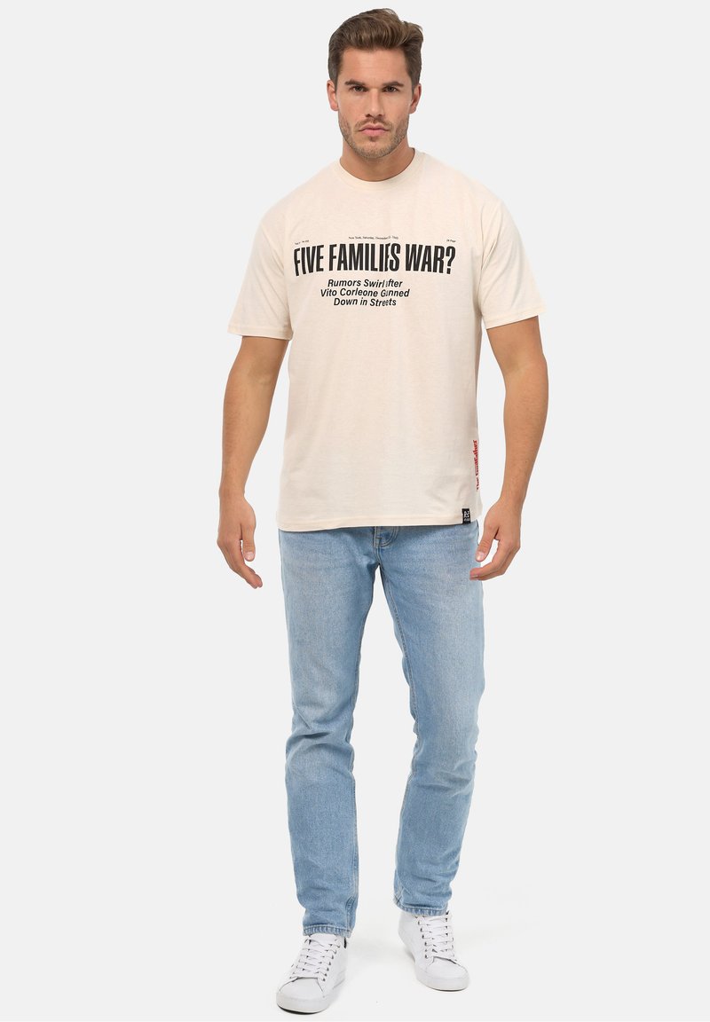 Camiseta de algodón beige con texto negro, cuello redondo y mangas cortas. Combinada con jeans azules claros y zapatillas blancas.