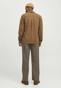 Brown, textured jacket with a collar, buttoned cuffs, and back yoke detail, paired with loose, light brown trousers and dark footwear.