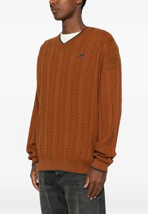 Uomo che indossa un maglione a punto treccia color ruggine con scollo a V e bordatura nera, e jeans scuri, in piedi davanti a uno sfondo semplice.