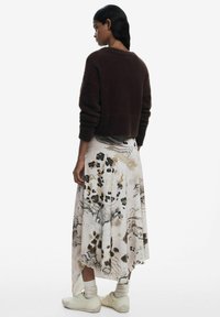 Maglione corto in peluche marrone, gonna lunga a fantasia con tonalità chiare e design astratti scuri, abbinata a scarpe chiare e calzini leggeri.
