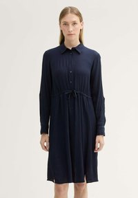 Abito blu navy, con bottoni, maniche lunghe, vita con fascia e spacchi laterali. Realizzato in tessuto leggero e testurizzato.