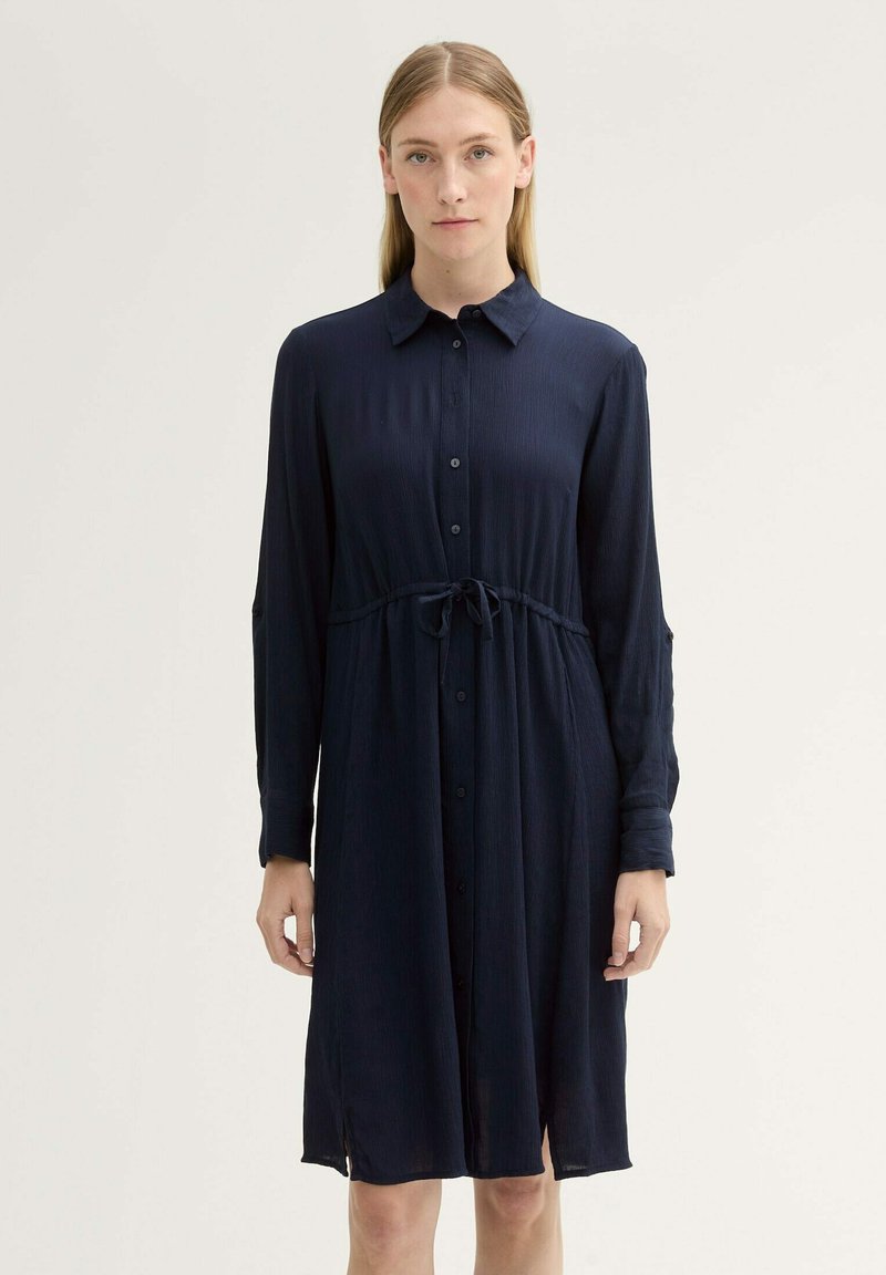 Abito blu navy, con bottoni, maniche lunghe, vita con fascia e spacchi laterali. Realizzato in tessuto leggero e testurizzato.