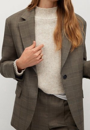 Femme portant un costume à carreaux marron avec un blazer à un bouton sur un pull en maille côtelée beige, montrant une main ornée de deux bagues en or.
