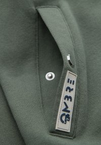 Gros plan sur un tissu vert avec un bouton-pression et une étiquette rectangulaire cousue indiquant le texte "N3R=" en bleu marine.