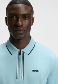 Ljust blå polo t-shirt med dragkedja i kragen, svarta detaljer och en 'BOSS' logotyp. Tillverkad av ett mjukt tyg med en subtil struktur.