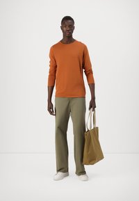 Oranges Longsleeve-Shirt, grüne Hose und weiße Sneaker. Trägt eine beige Tragetasche. Einfaches Design und lockere Passform.