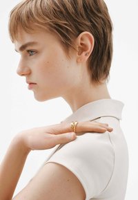 Pendientes de oro con una cadena larga y delicada y un anillo de oro anudado. El sujeto lleva una camisa de color claro con cuello. Textura suave.