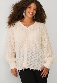 Vrouw met krullend haar die een textuurrijke crèmekleurige blouse met lange mouwen, zwarte broek en hangende kralenoorbellen draagt, lachend naar de camera.