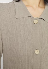 Cardigan bege canelado com colarinho, apresentando dois botões redondos de madeira e uma textura suave, enfatizando o padrão de linhas verticais.