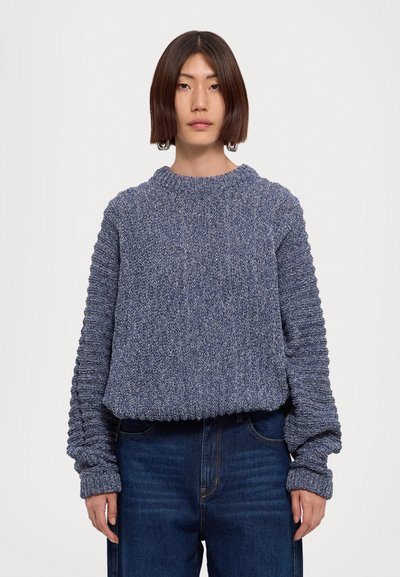Suéter de punto texturizado en color navy con puños y dobladillo acanalados. Escote redondo, corte relajado y un patrón intrincado en las mangas. Combinado con jeans oscuros.