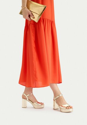 Kobieta w jasnopomarańczowej sukience midi trzyma błyszczącą złotą kopertówkę i nosi pasujące do nich złote sandały na platformie z paskami wokół kostek.