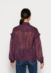 Blusa floral com tecido roxo transparente e detalhes em rosa, gola alta e mangas bufantes. O design das costas inclui detalhes franzidos.