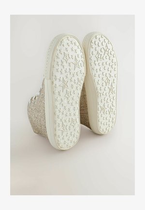 Spīdīgi zelta platformas sporta apavi ar baltiem gumijas zolēm, uz kurām ir zvaigžņu raksts, metāla caurumiņiem un teksturētu virsmu.