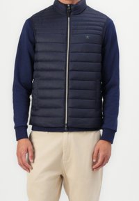 Homme portant un gilet matelassé sans manches bleu marine sur une chemise à manches longues bleu marine et un pantalon beige, sur fond blanc.