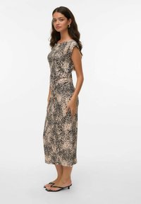 Femme portant une robe midi sans manches beige et noire à pois, avec des sandales à talons noirs, se tenant devant un fond blanc uni.