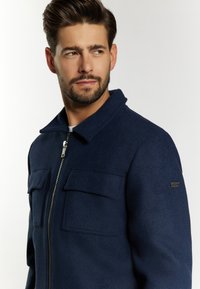 Dunkelblaues Wollmischjackett mit Reißverschluss, spitzem Kragen und zwei Brusttaschen mit Klappen. Dezentes Logo-Akzent am Ärmel.