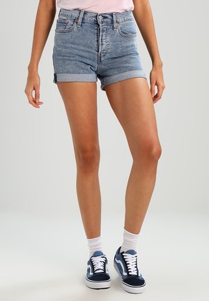 Person iført opsmøgede denimshorts, hvide sokker og blå sneakers, stående mod en ensfarvet lys baggrund.