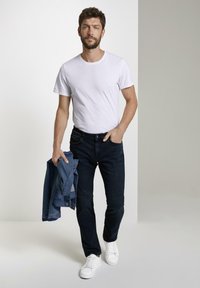 Bílá tričko, tmavě modré džíny a bílé tenisky. Drží modrou džínovou bundu. Pohodlný střih a jednobarevné bez viditelných vzorů.