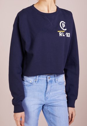 Femme portant un sweat-shirt court bleu marine avec le logo "PC RL-93" et un jean taille haute bleu clair, debout devant un fond uni.