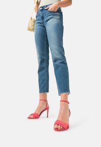 Žena v krátkých modrých džínách a růžových vysokých podpatcích, držící malou béžovou kabelku, stojící na jednolitém pozadí.