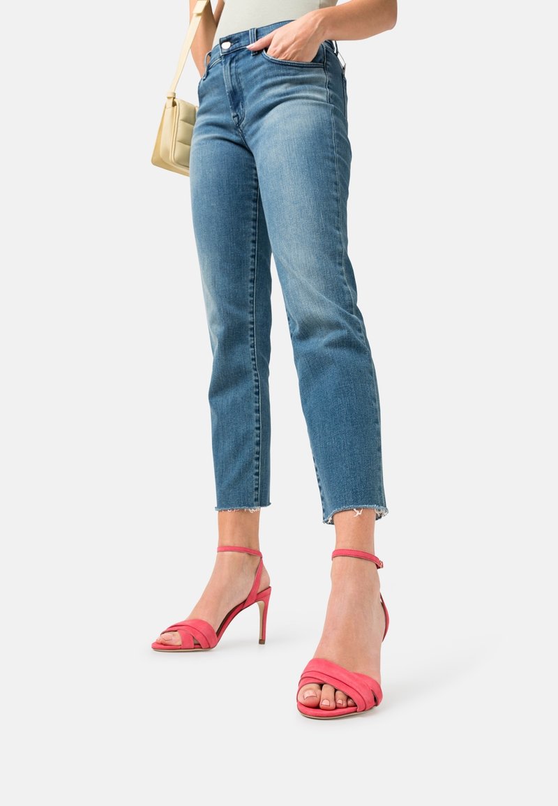 Žena v krátkých modrých džínách a růžových vysokých podpatcích, držící malou béžovou kabelku, stojící na jednolitém pozadí.