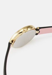 Guldtonad klocka med ett slätt silverhölje, svart läderrem och rosa läderdetalj. Har en rund form och en krona för justering.