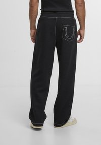 Pantalones de chándal negros con detalles de costura blanca, corte relajado, cintura elástica y un bolsillo trasero con un diseño de logotipo distintivo.