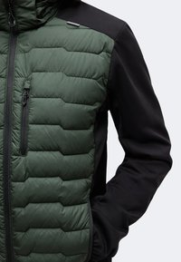 Veste matelassée verte devant avec manches noires, dotée d'une poche poitrine zippée verticale et d'une fermeture éclair frontale complète.