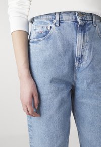 Persona vistiendo jeans de mezclilla azul claro y una blusa blanca de manga larga, con la mano izquierda relajada a su lado.